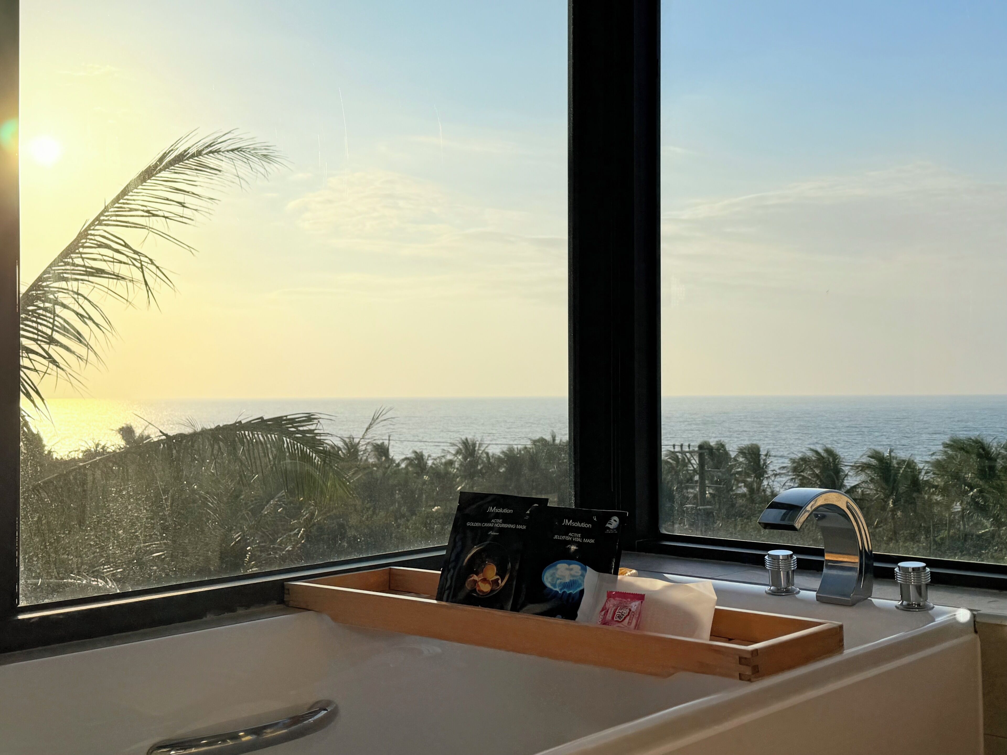 A bathroom with a view of the ocean, a wooden tray with toiletries, and a window with a view of palm trees and the sea.