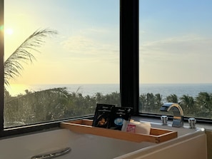 A bathroom with a view of the ocean, a wooden tray with toiletries, and a window with a view of palm trees and the sea.