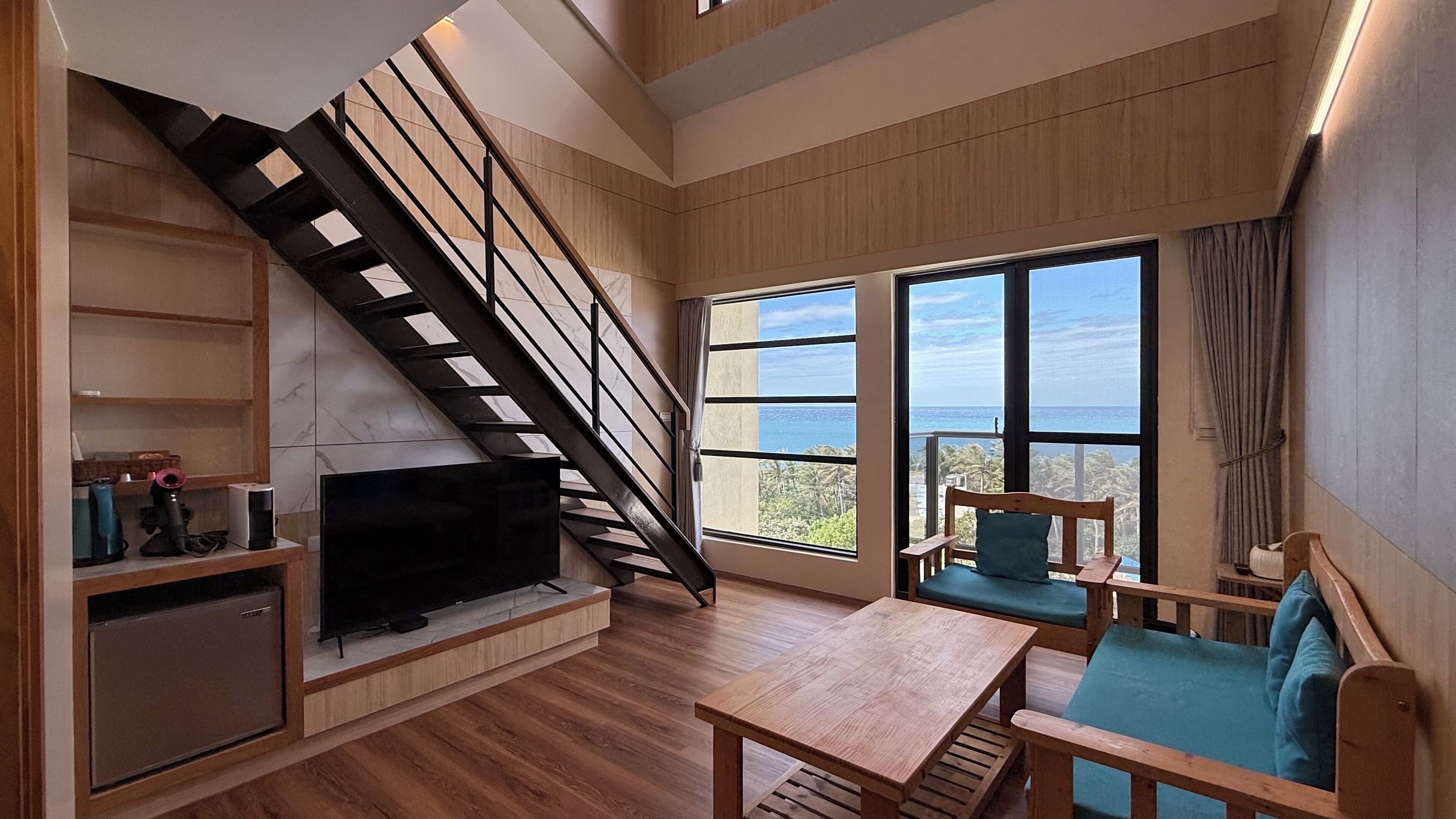 A modern living room with a wooden staircase, a flat-screen TV, a dining table, and a sofa with a view of the ocean.