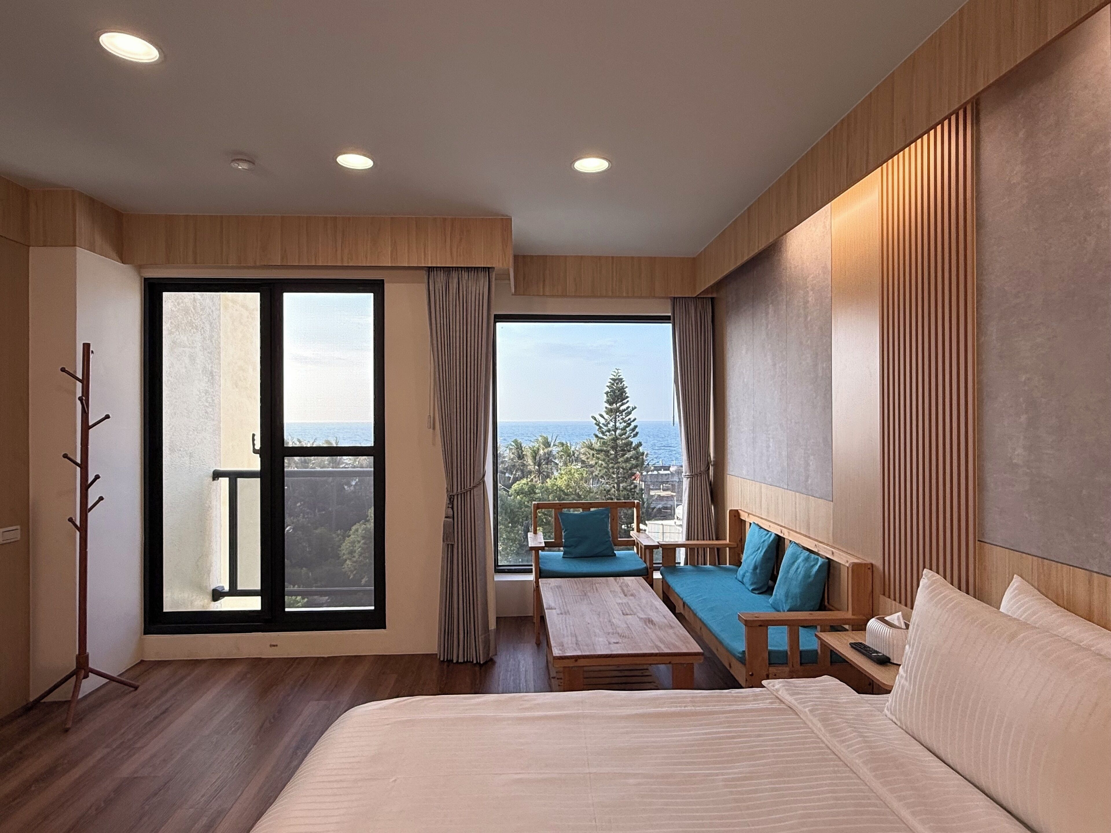 A modern hotel room with a large bed, a sofa, a table, and a balcony with a view of the sea.