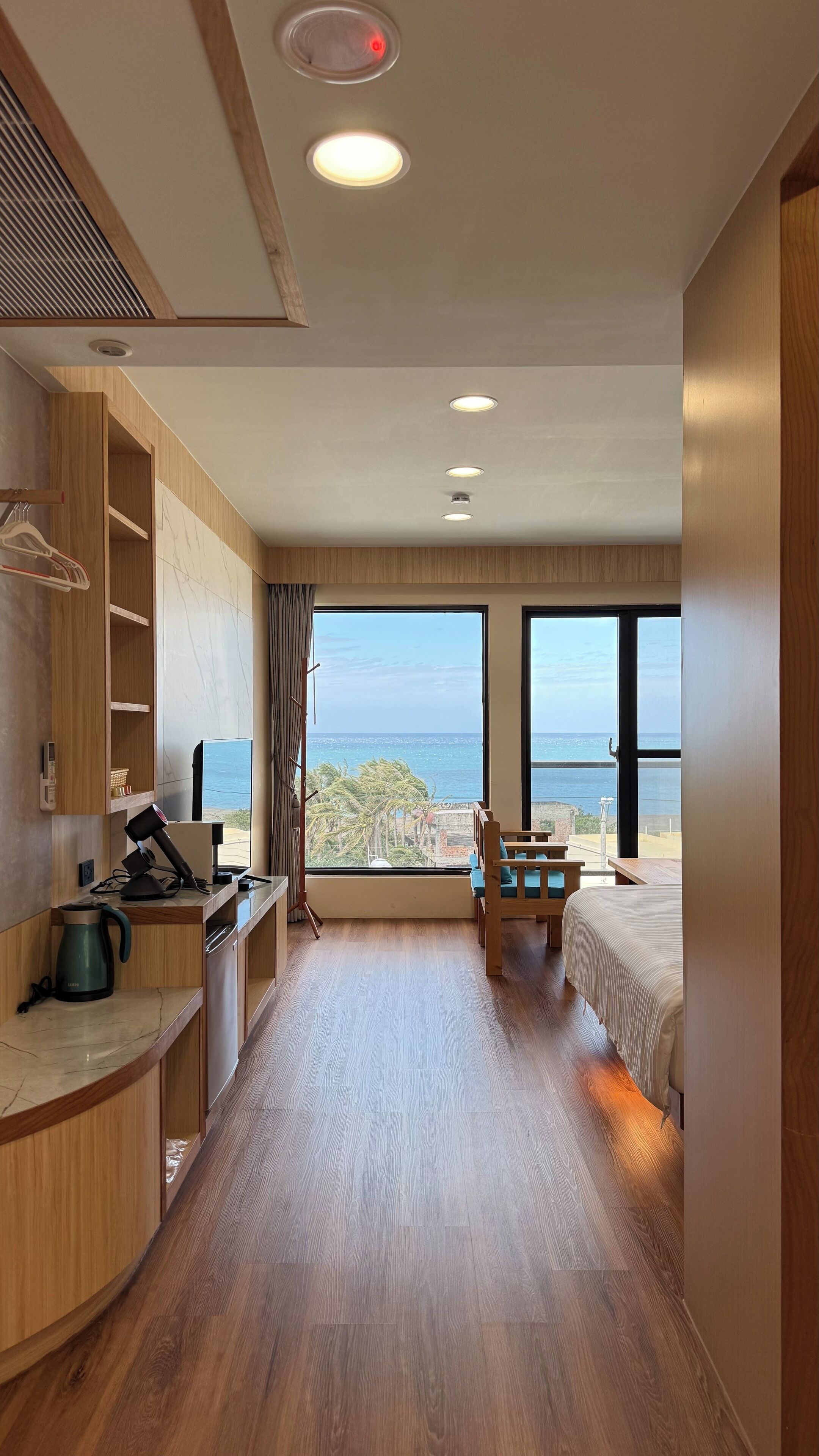 A modern hotel room with a wooden floor, a bed, a desk with a kettle, a chair, and a view of the sea.