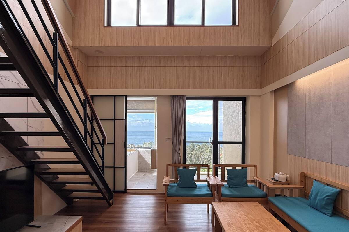 A modern interior with a wooden staircase, a seating area with blue cushions, and a view of the sea through large windows.