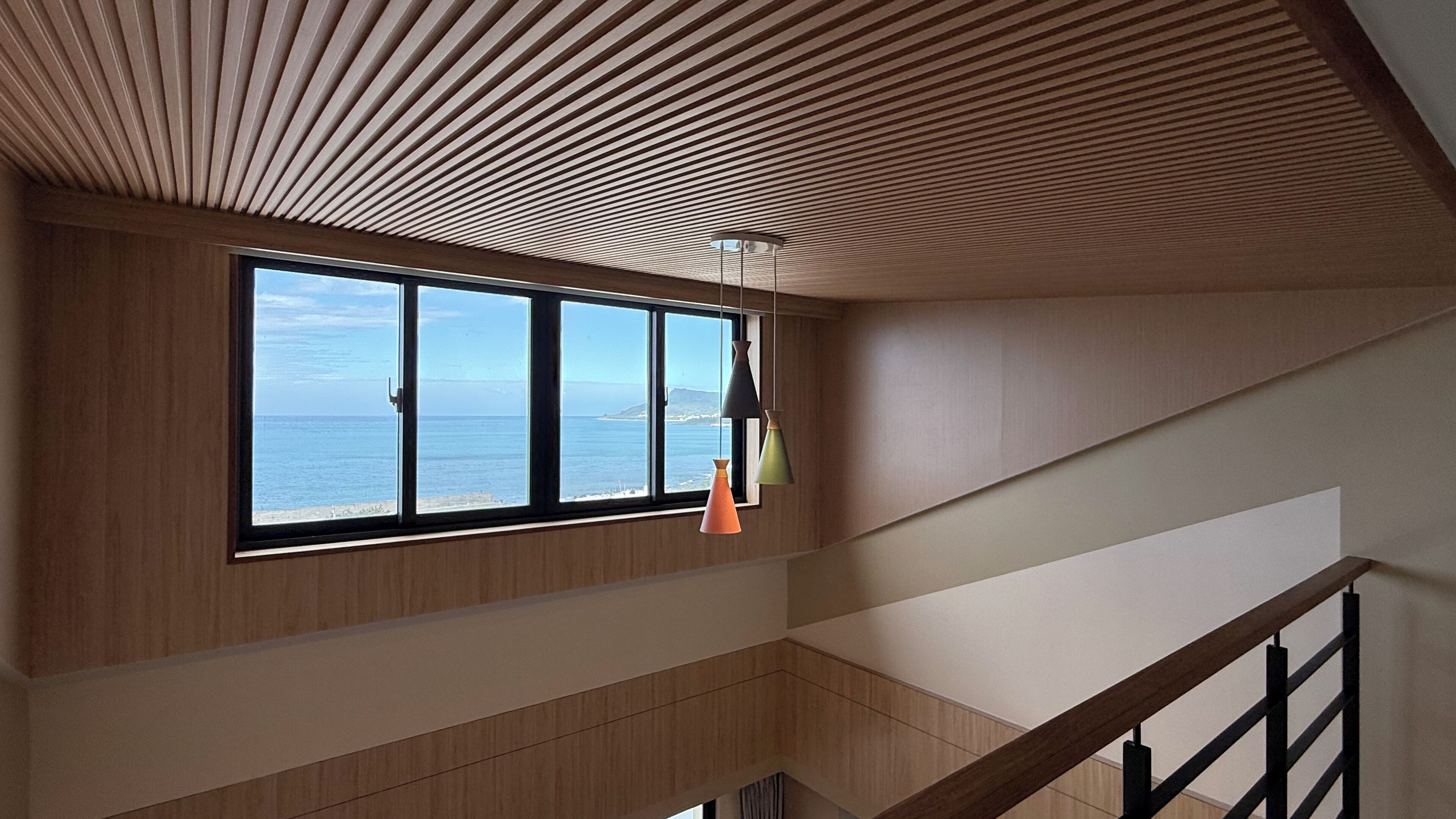 Un interior moderno con techo inclinado, un ventanal grande con vistas al mar y una escalera con barandilla de madera.