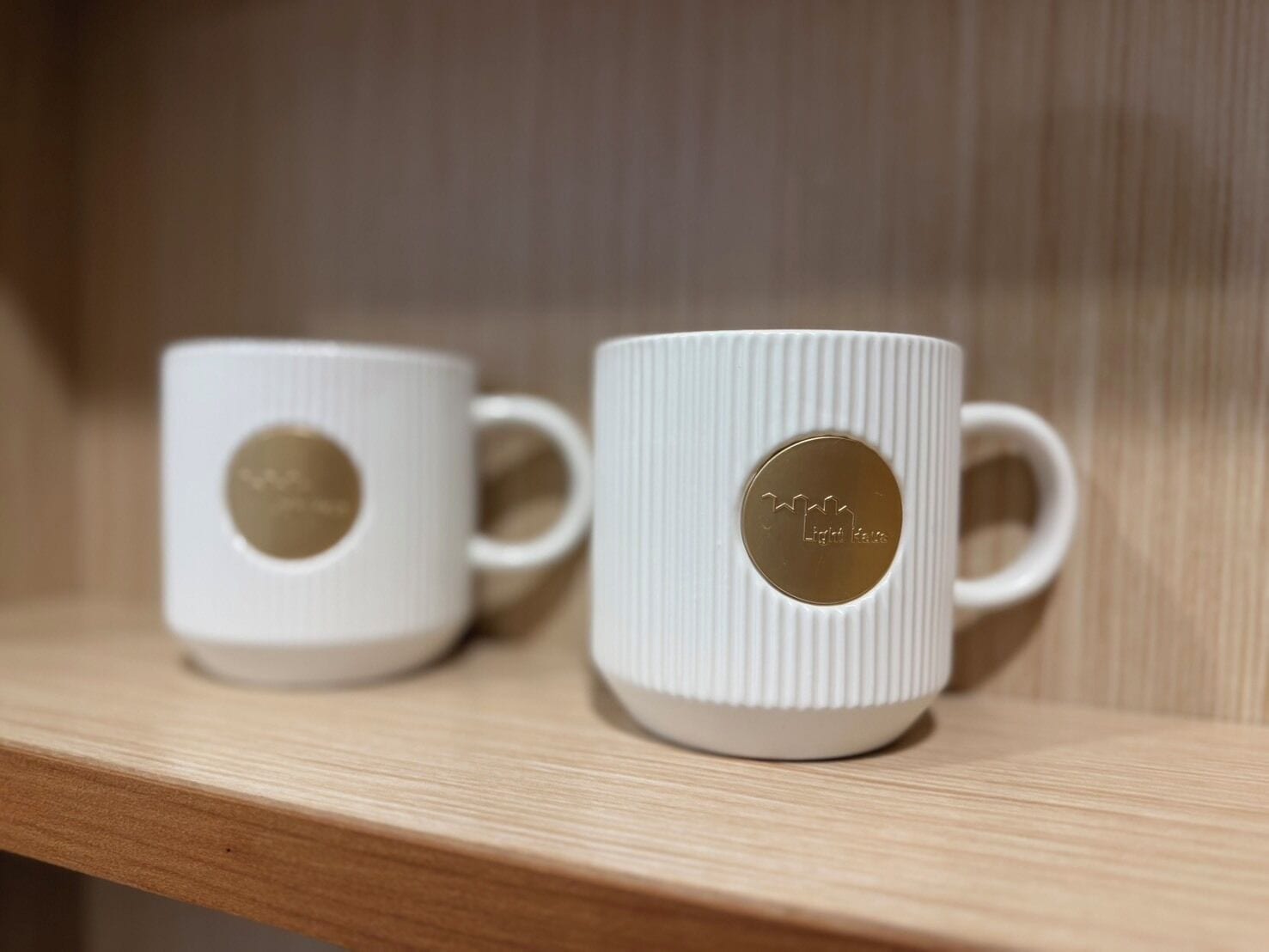 Two white ceramic mugs with embossed logos on a wooden shelf.