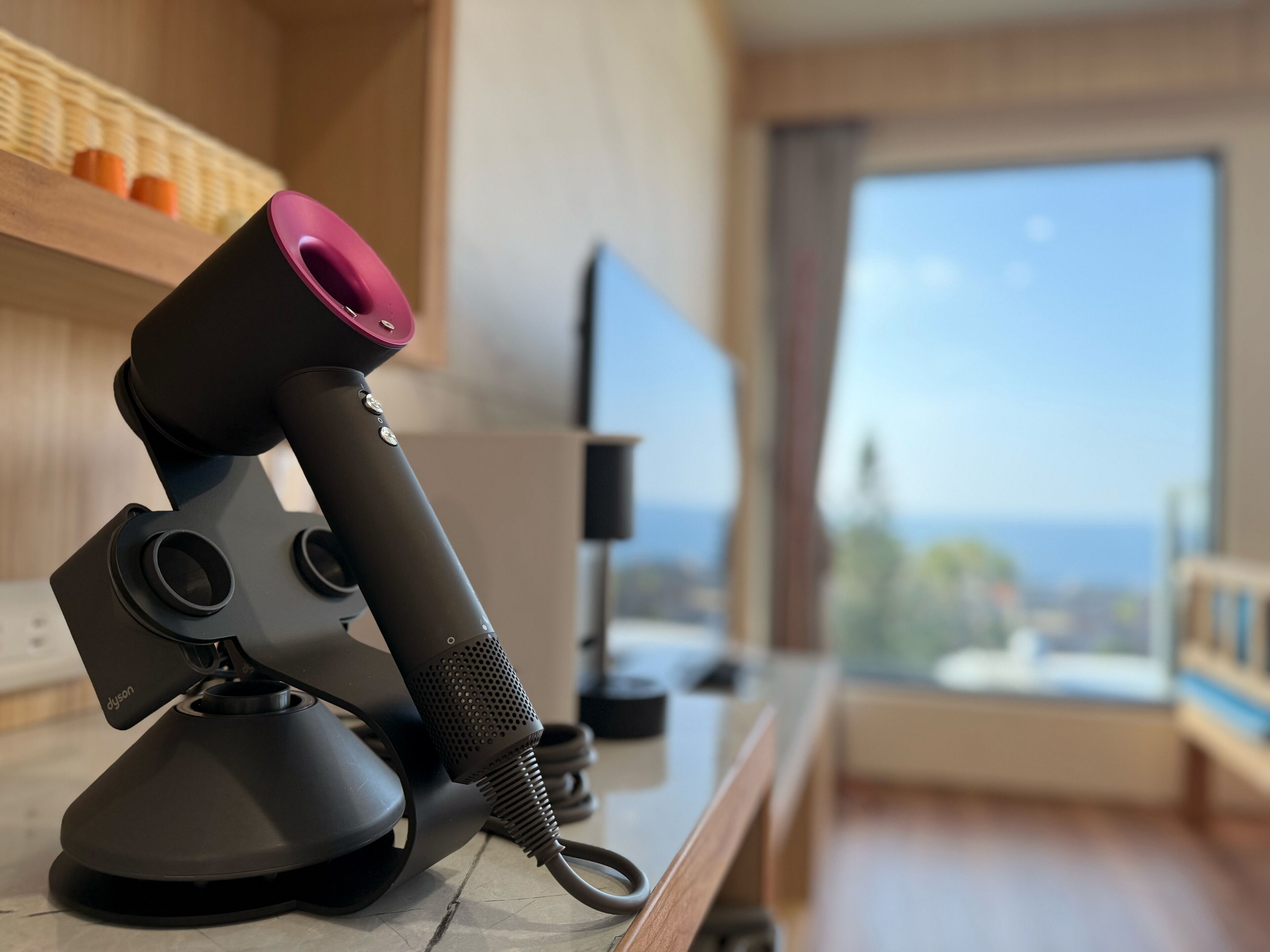 Un secador de pelo Dyson negro, moderno y compacto, sobre una encimera de madera, con vistas a un paisaje urbano a través de un ventanal amplio.