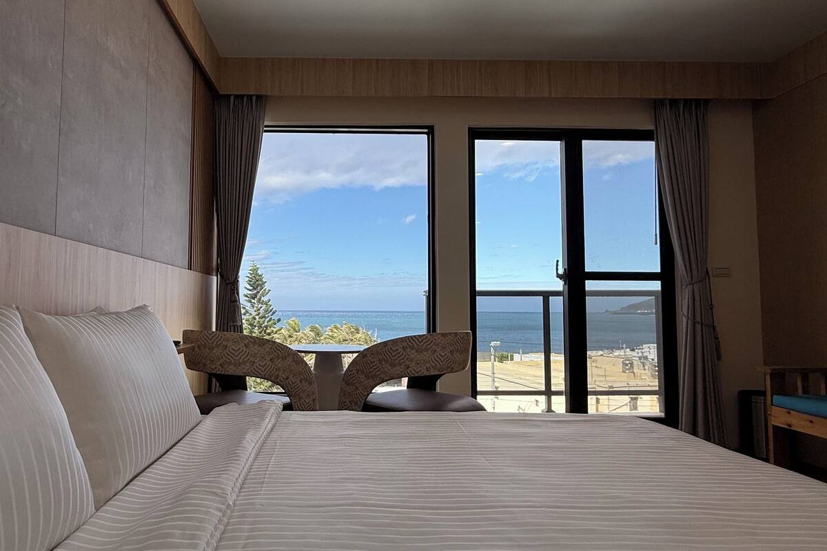 A hotel room with a large bed, two chairs, a view of the sea, and a balcony.