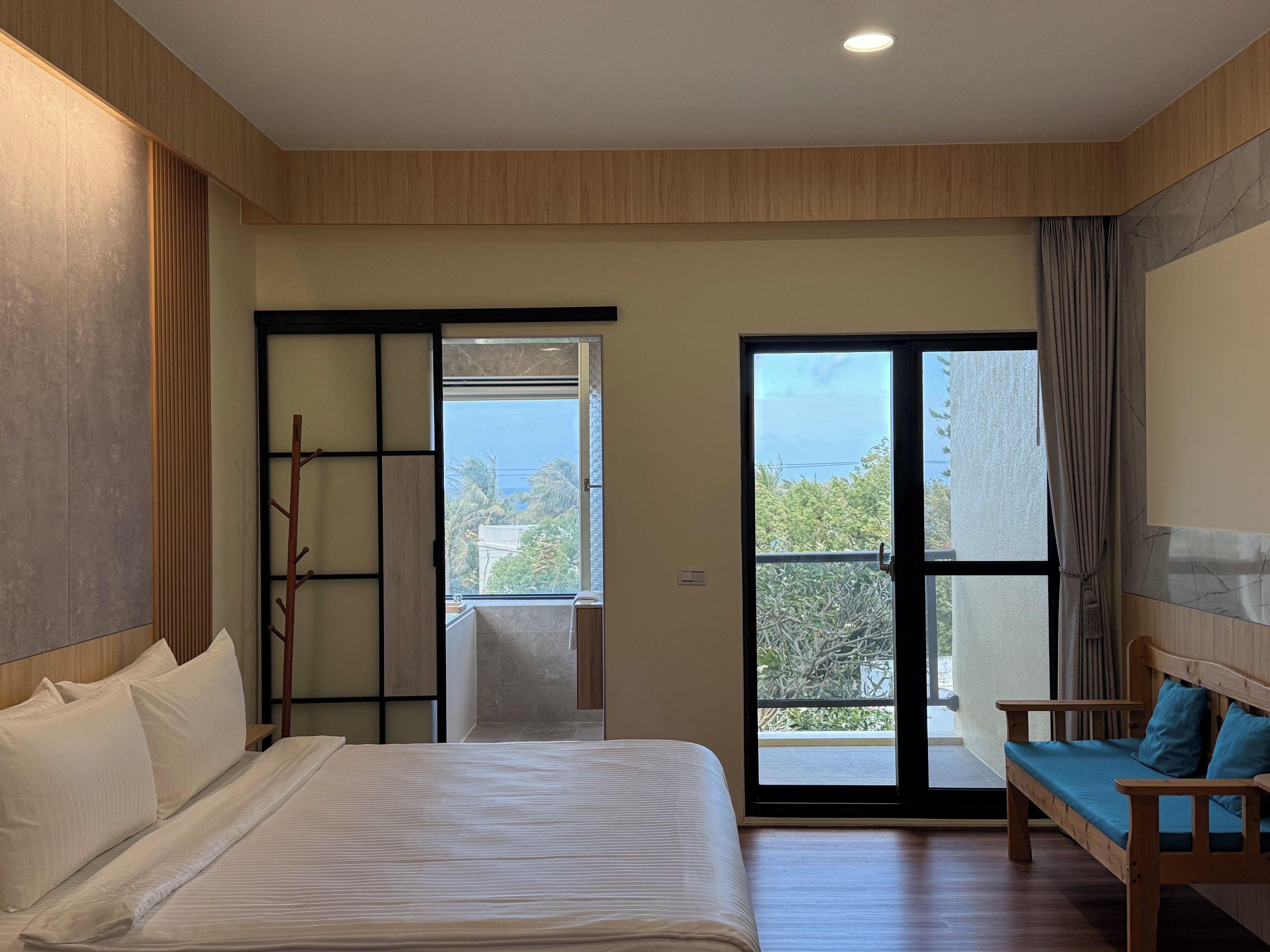 Un dormitorio moderno con cama, un banco de madera y un ventanal corredera que da a un balcón.