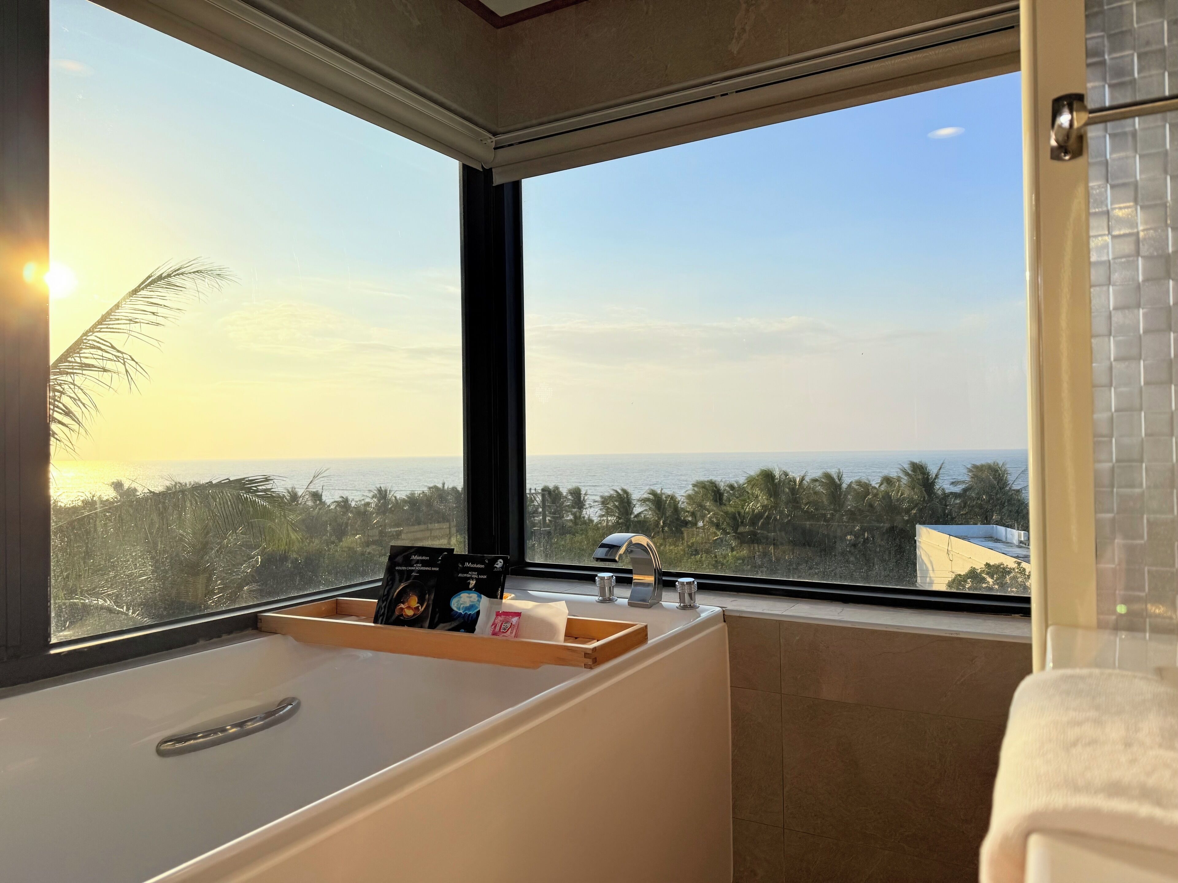 A bathroom with a bathtub, a window with a view of the sea and palm trees, and a towel rack.