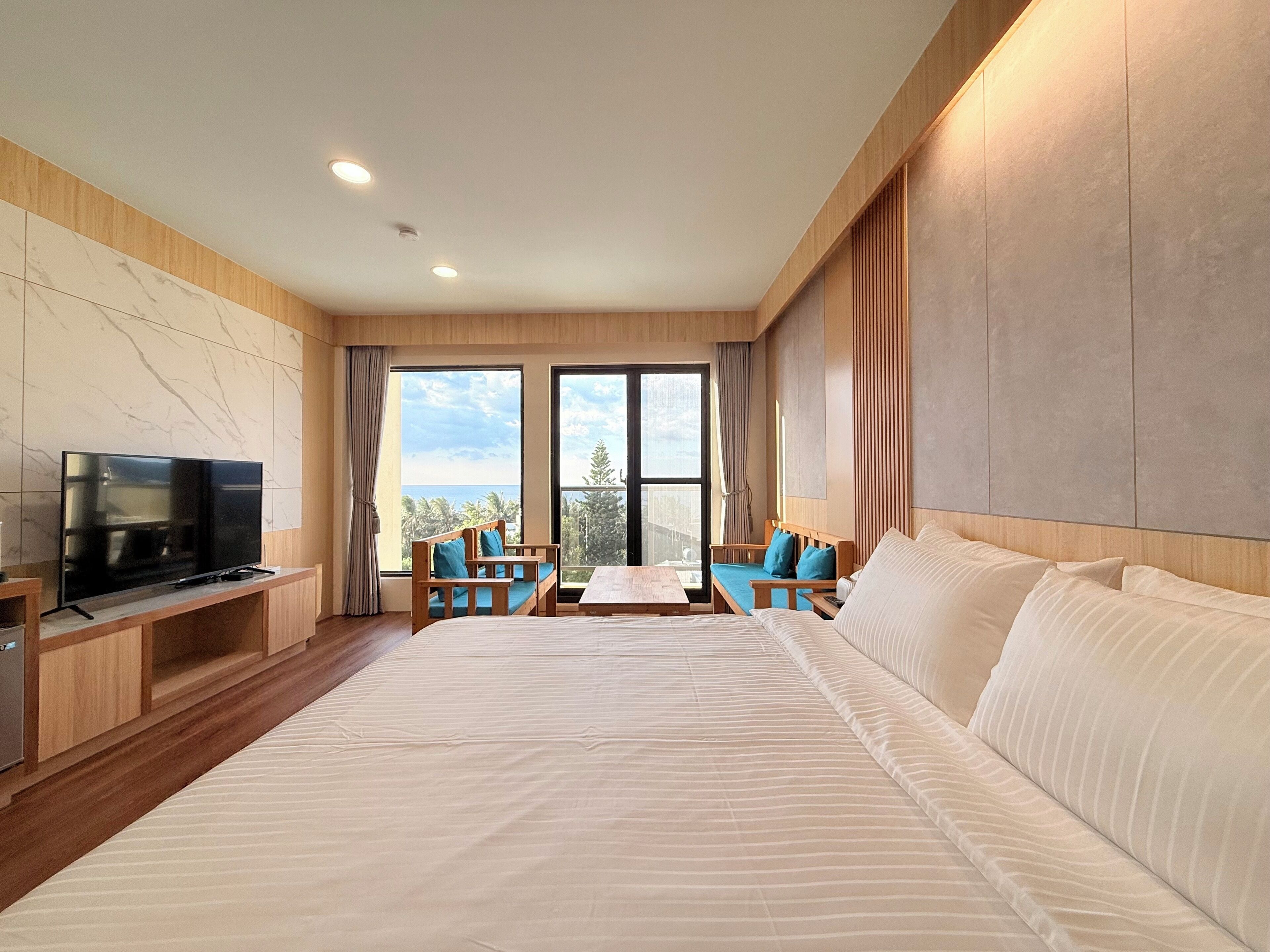 Een moderne hotelkamer met een groot bed, een televisie op een houten standaard, een eettafel met stoelen en grote ramen met uitzicht op het omliggende landschap.