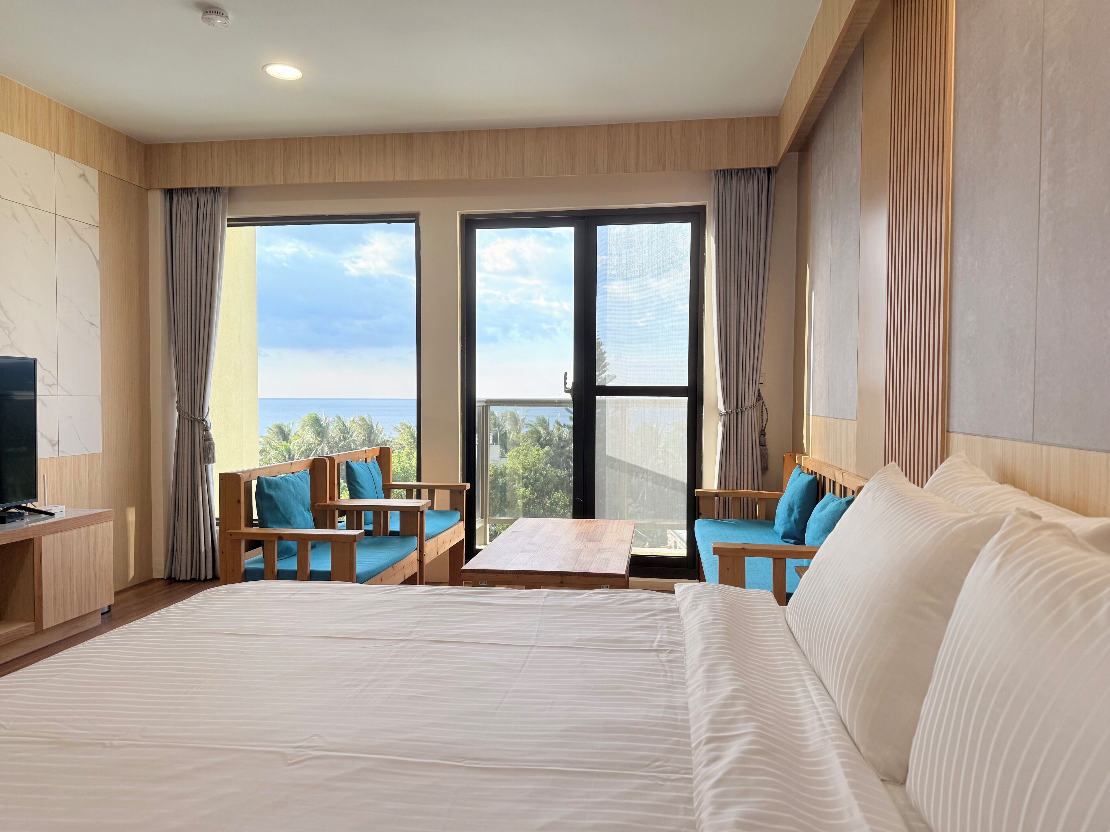Ein Hotelzimmer mit einem großen Bett, zwei Holzstühlen mit blauen Kissen, einem kleinen Tisch und einem Fernseher auf einer Holzkommode.