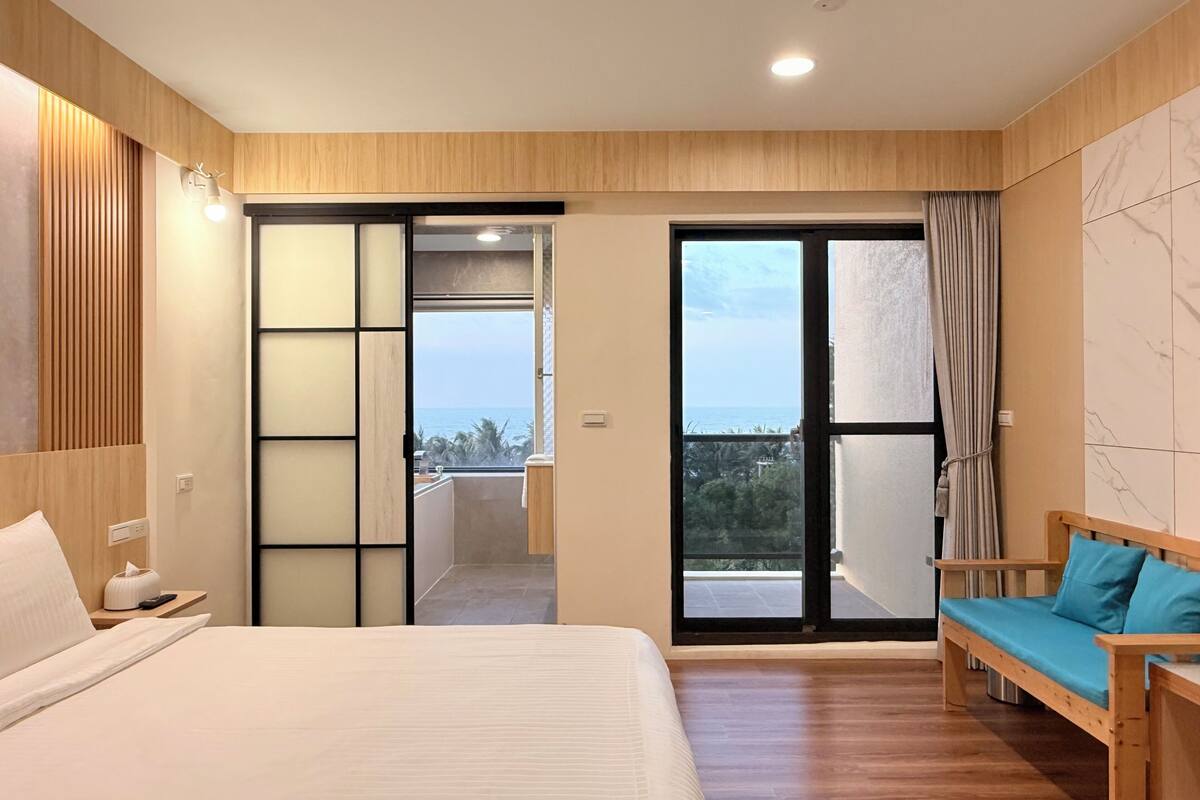 A hotel room with a bed, a wooden bench, a sliding door to a balcony, and a view of the sea.