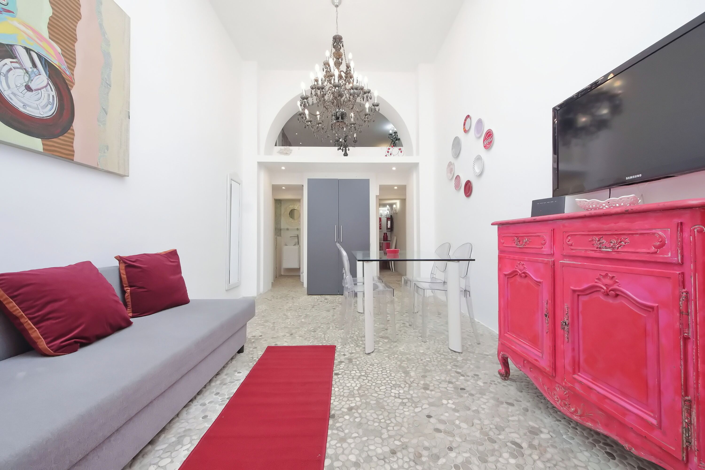 Una sala de estar con un sofá gris, cojines rojos, un tapete rojo, un mueble rosa y un televisor sobre un soporte.
