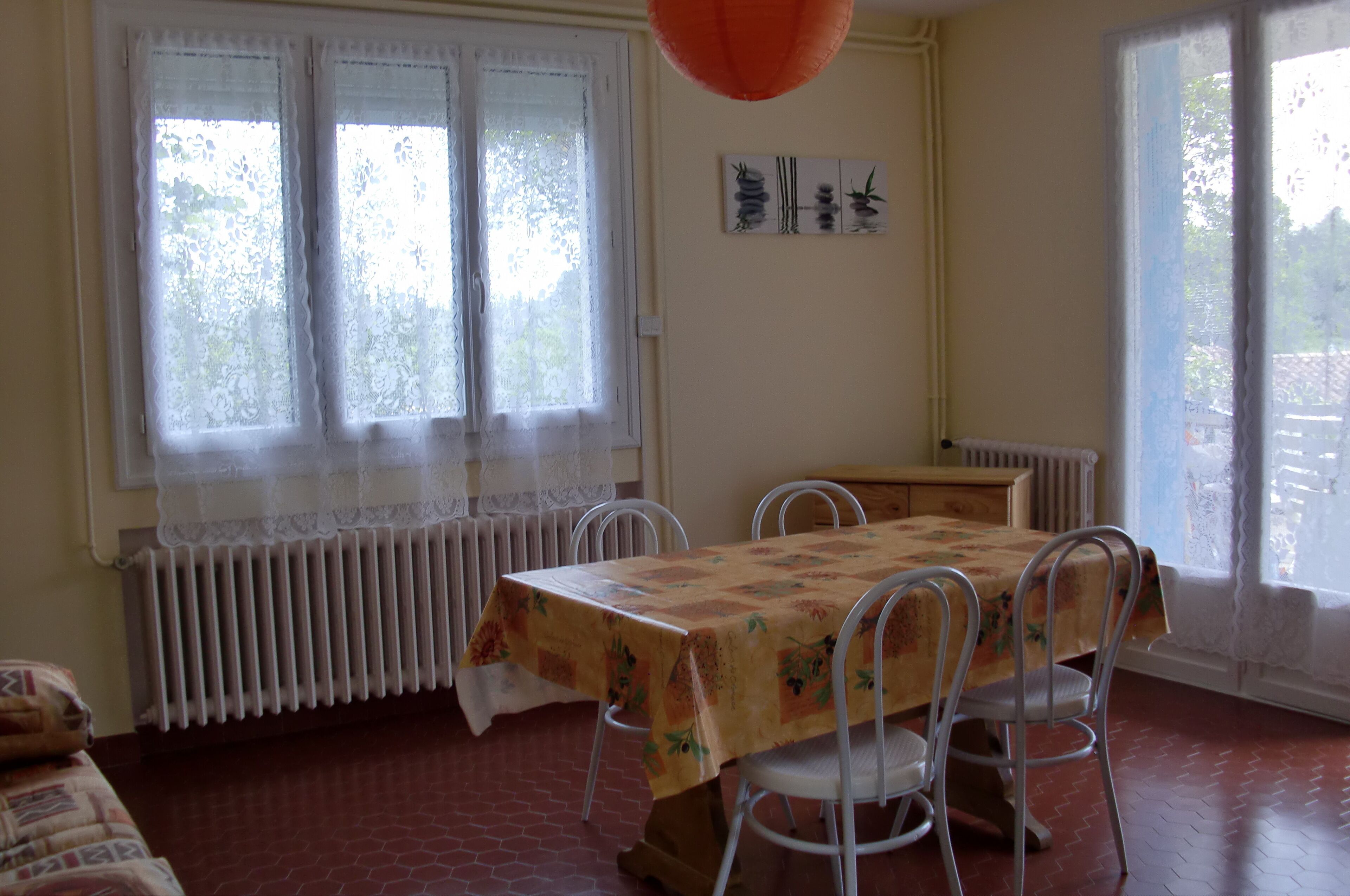 Et spiseområde med et bord dækket af en blomstret dug, hvide stole, en radiator, blondegardiner og et vindue med udsigt til grønne områder.