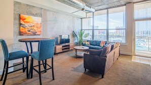 Een moderne woonkamer met een bank, stoelen, een salontafel en een televisie. Er hangt een schilderij aan de muur en een groot raam met uitzicht op de stad.