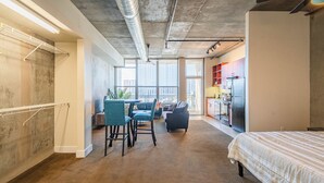 Een moderne hotelkamer met een groot bed, een zithoek met stoelen, een kitchenette en uitzicht op de stad door grote ramen.