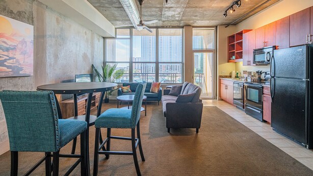 Een moderne keuken met een zwarte koelkast, magnetron en fornuis. Er is een eethoek met een ronde tafel en stoelen. De kamer heeft grote ramen met uitzicht op de stad.