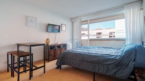 Ein Schlafzimmer mit einem Bett, einem Schreibtisch, einem Stuhl, einem Fernseher und einem Fenster mit Blick auf Gebäude.