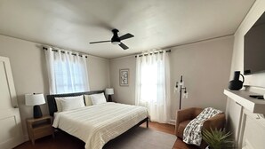 Un dormitorio con cama, mesitas de noche, ventilador de techo, lámpara de pie, una silla, planta en maceta y chimenea.