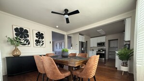 Un comedor con una mesa de madera y sillas acolchadas, un ventilador de techo y vista a la cocina.