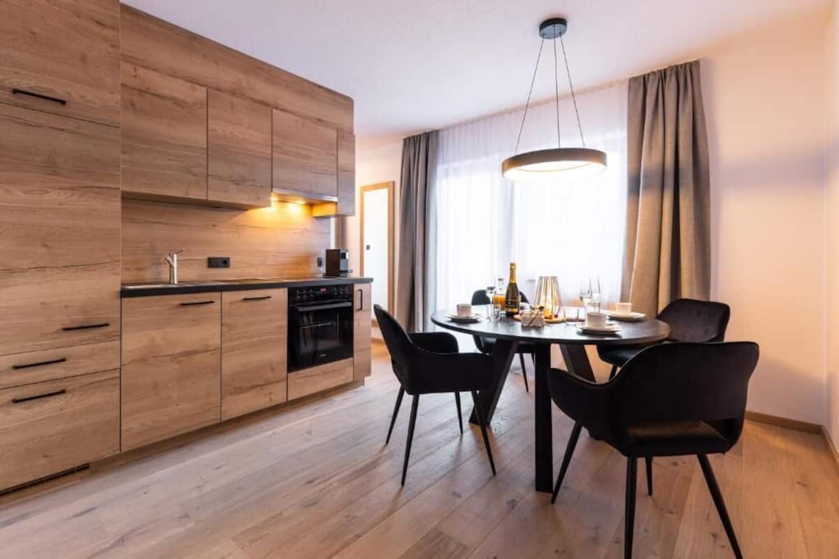 A modern kitchen with wooden cabinets, a dining table with chairs, and a large window with curtains.