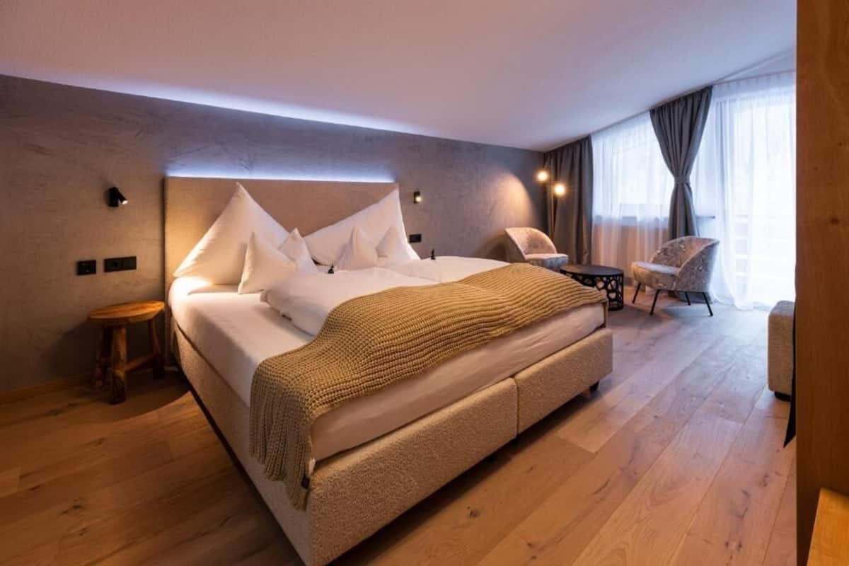 A modern hotel room with a large bed, a small side table, a chair, and a view of the outdoors through large windows.