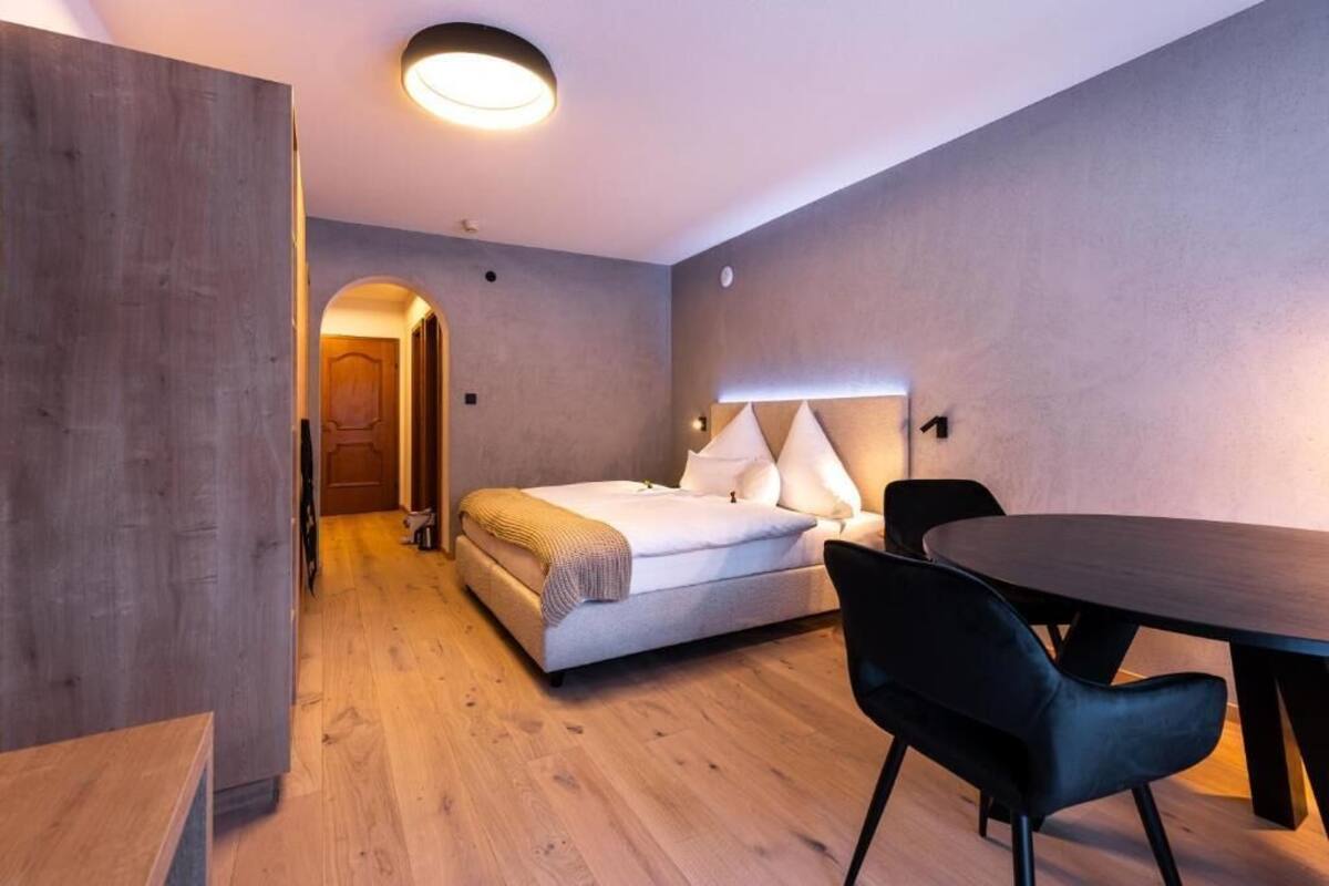 A modern hotel room with a wooden floor, a bed with white linens, a round dining table, and a black chair.