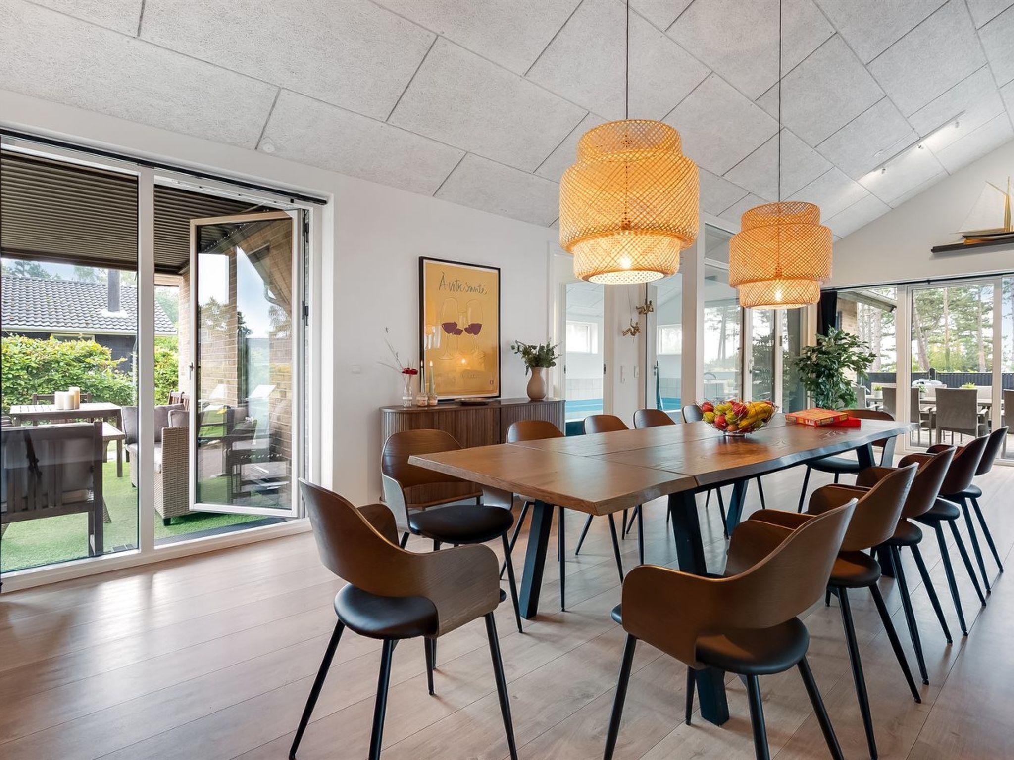 Un vaste espace dédié à la salle à manger, avec une grande table en bois et des chaises modernes, de grandes fenêtres et deux lampes suspendues en osier.