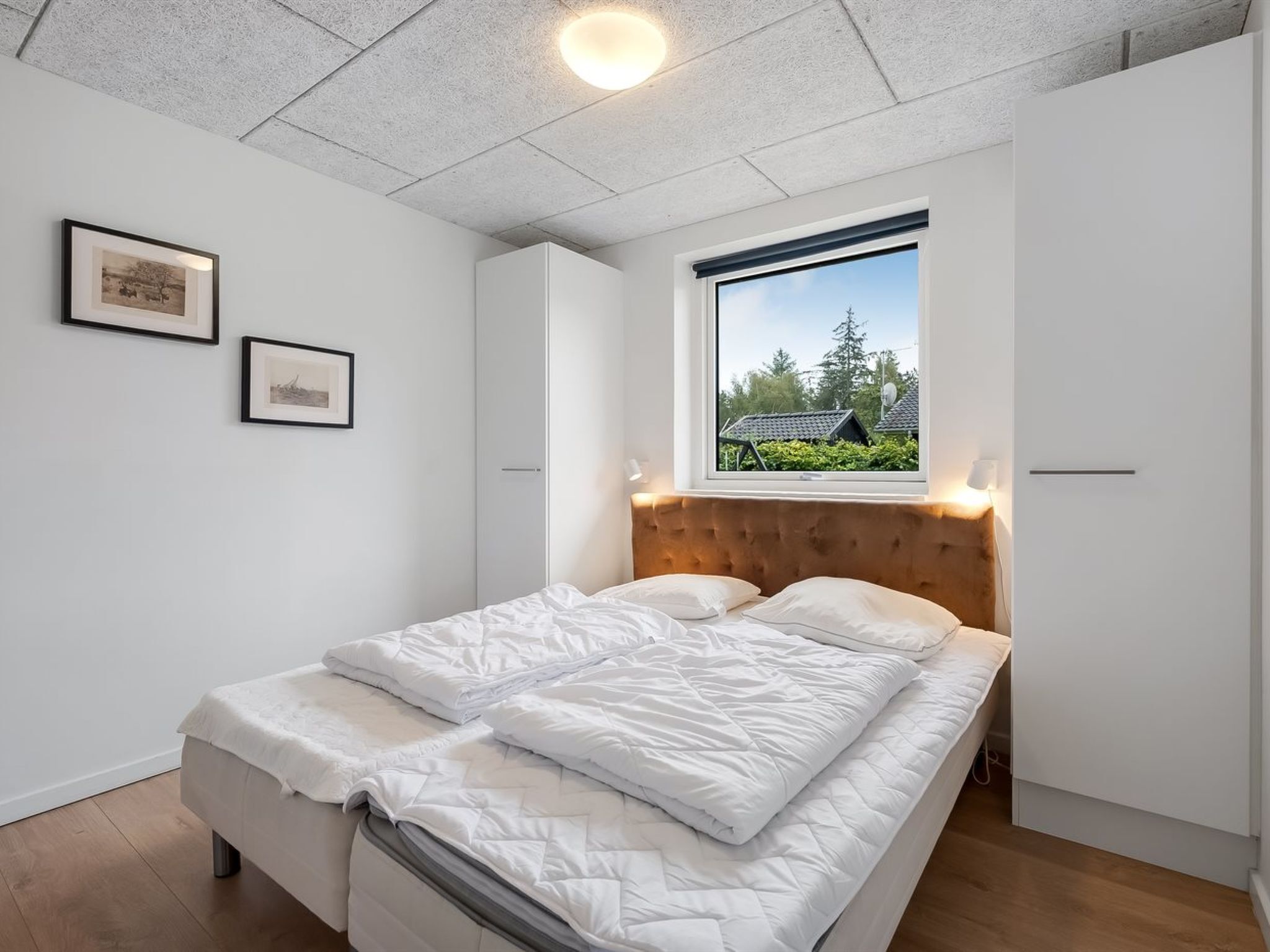 Une chambre à coucher avec un lit, une fenêtre donnant sur des maisons et deux tableaux encadrés accrochés au mur.