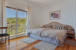 Een slaapkamer met een groot bed, een nachtkastje, een stoel, een balkon met uitzicht op groen, en een lijst met een afbeelding aan de muur.