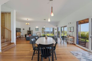 Een ruime woonkamer met een eettafel, stoelen, een bank en een televisie. Links bevindt zich een trap en er is uitzicht op het buitenland via grote ramen.