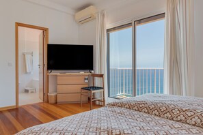 A hotel room with a bed, a television, a desk with a chair, a small stool, and a window with a view of the sea. - Sea Sky House By Holiday Rental Madeira (Ribeira Brava)