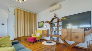 Una sala de estar con un sofá, un sillón amarillo, una mesa de centro redonda, una estantería y un televisor de pantalla plana.