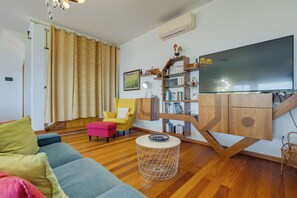 Una sala de estar con un sofá, un sillón amarillo, una mesa de centro redonda, una estantería y un televisor de pantalla plana.
