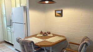 Une petite cuisine avec un réfrigérateur blanc, une table en bois et deux chaises, ainsi qu’un luminaire suspendu.