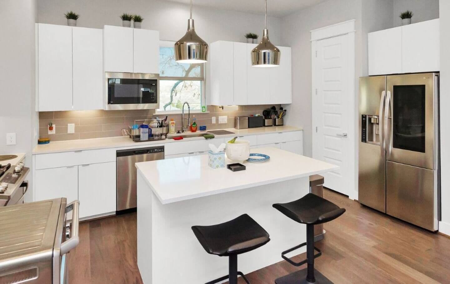 A modern kitchen with white cabinetry, stainless steel appliances, a central island with stools, and pendant lighting.
