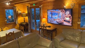 Un acogedor interior de cabaña con paredes de madera, una gran pantalla de televisión mostrando una escena festiva, sofás de cuero y un comedor con mesa y sillas.