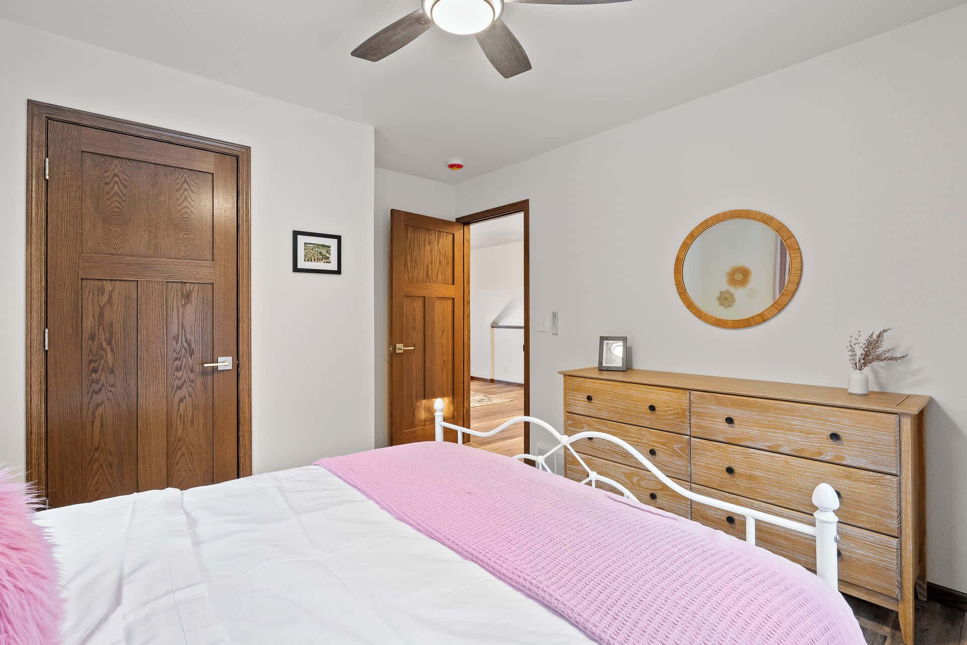 Ein Schlafzimmer mit Bett, Schminktisch, Holztür, Deckenventilator und einem gerahmten Bild an der Wand.
