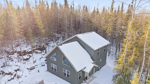 Une maison à deux étages, dotée d’un toit à forte pente, entourée d’une zone boisée où la neige recouvre le sol.