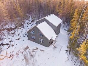 A two-story house with a snow-covered roof, surrounded by a forested area.