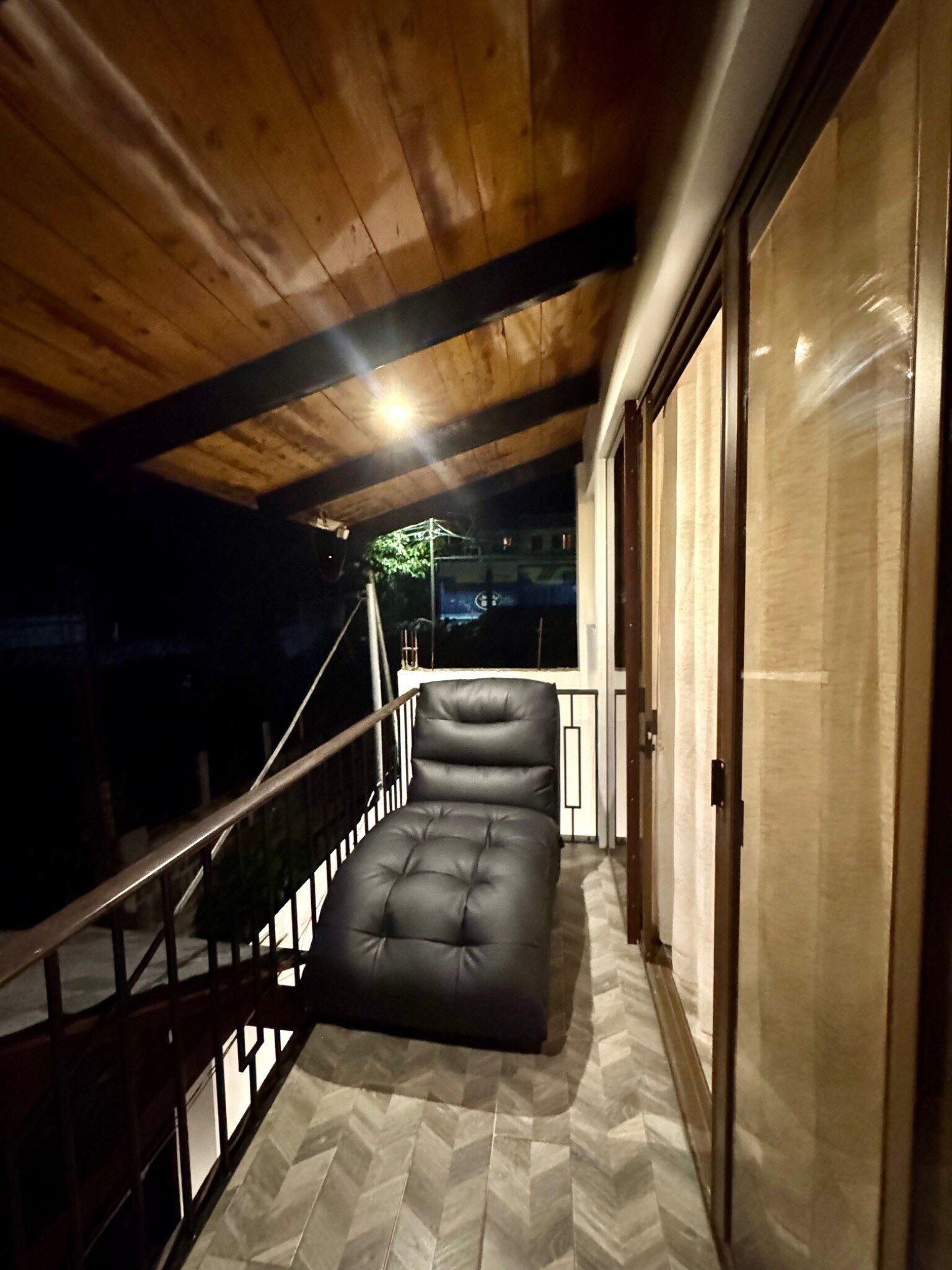 Un balcón con una zona de estar acolchada, vigas de madera en el techo y un ventanal de vidrio que da a un espacio interior.