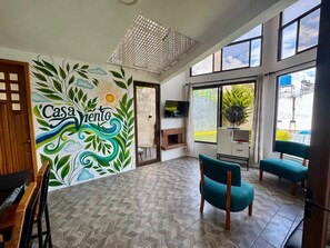 A spacious room with a herringbone floor, a large window with a view of greenery, and a mural with the word "Casa" and a sun. - Casa Viento - Private Pool, Garden & BBQ (San Pedro La Laguna)