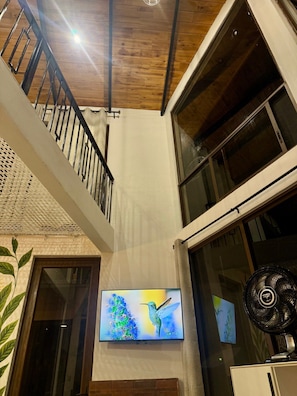 Un intérieur moderne avec un plafond en bois, un escalier doté de rampes en métal, un téléviseur à écran plat fixé au mur et un ventilateur sur pied.