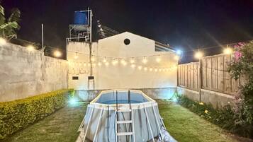 Una piscina exterior con escalera, rodeada de un césped cuidado y luces colgantes.