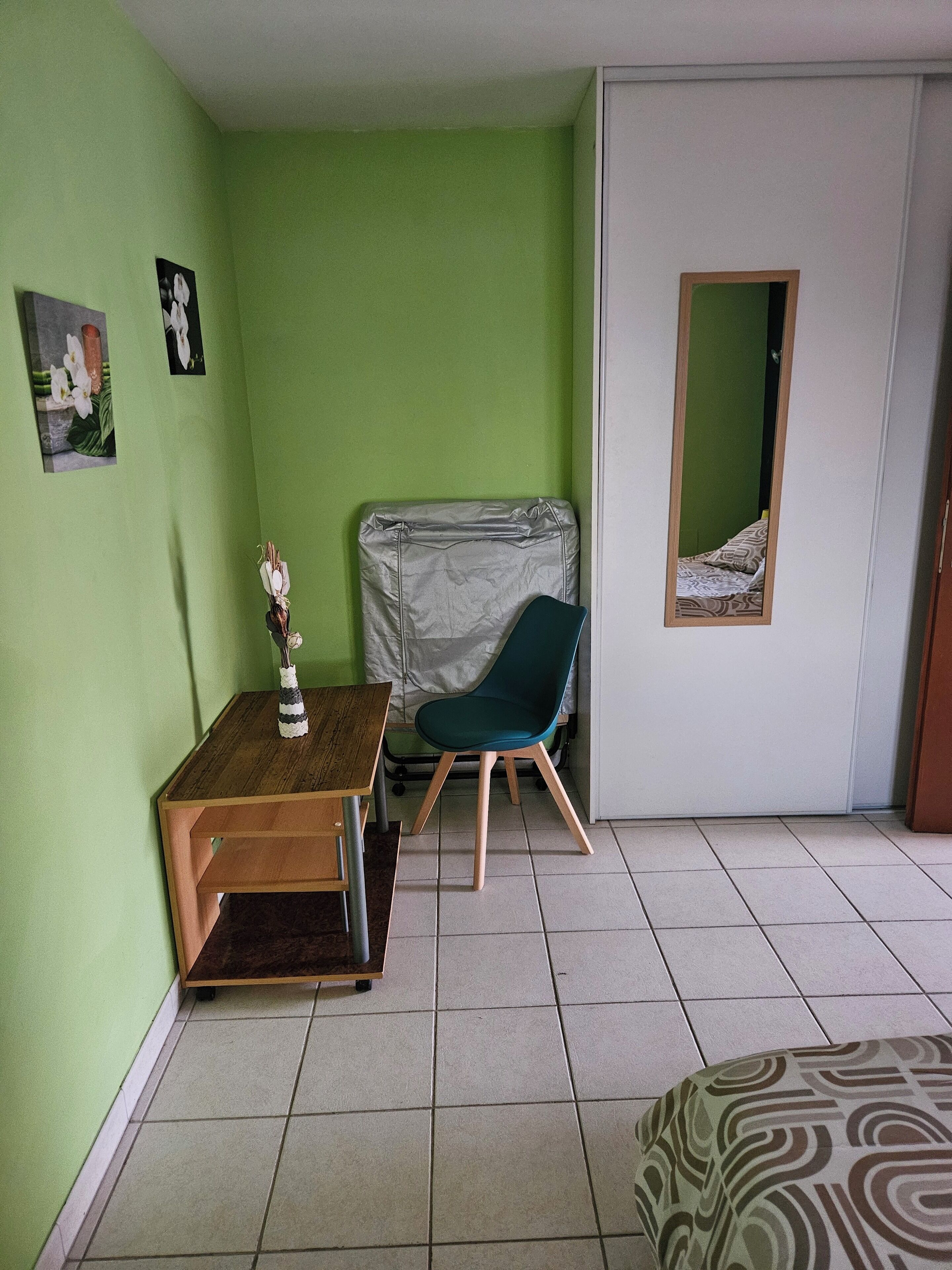 Une petite pièce aux murs verts, avec une table en bois, une chaise turquoise, un lit recouvert d’une literie à motifs et un miroir accroché au mur.