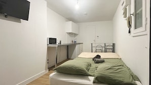 Una habitación pequeña y funcional con una cama individual, un escritorio con computadora, un microondas y una pequeña kitchenette.