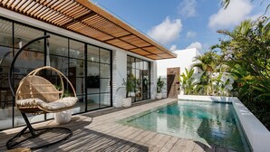 Un’area piscina moderna all'aperto con un solarium in legno, una poltrona in vimini e una stanza con ampie vetrate.