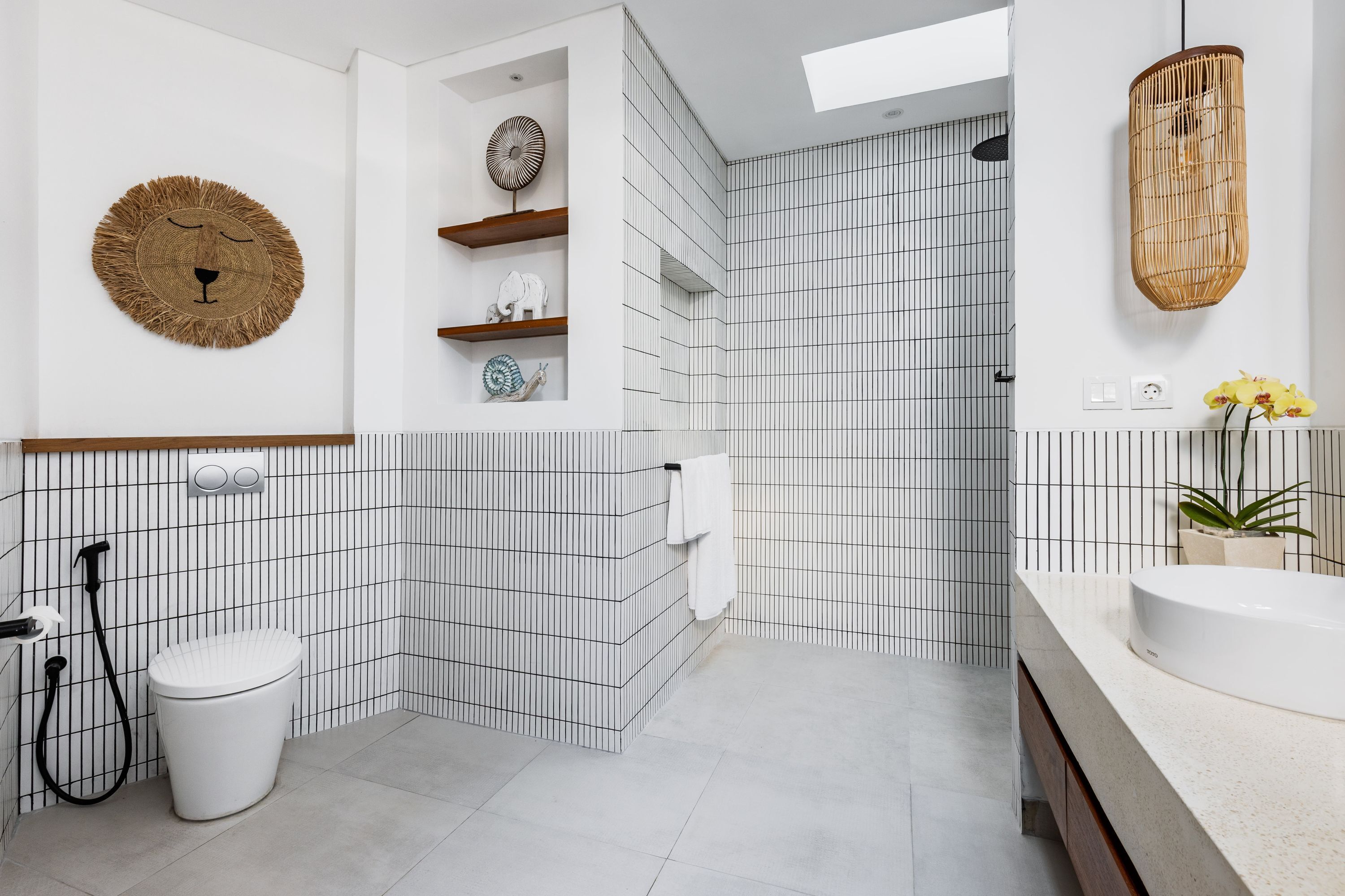 A modern bathroom with a white and black tiled wall, a white toilet, a shower area, and a vanity with a sink and a potted plant.