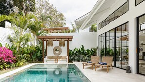 Ett modernt utomhusområde med pool, en pergola, solstolar och frodiga, tropiska växter.