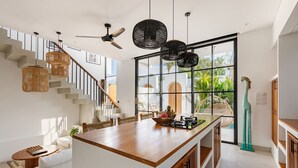A modern kitchen with a central island, a dining area with wicker furniture, and a staircase with a ceiling fan. - Family Nest 2BR Villa with pool and balcony (Kabupaten Badung)