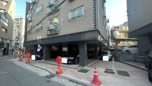A multi-story building with a parking garage entrance, several parked cars, and red traffic cones on the sidewalk. - Anne House (Seoul)